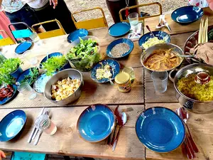 Zu sehen ist ein gedeckter Tisch von oben. Blaue Teller und Besteck stehen bereit sowie Schüsseln mit verschiedenen Gerichten, Dips, Salaten und Körnern. 