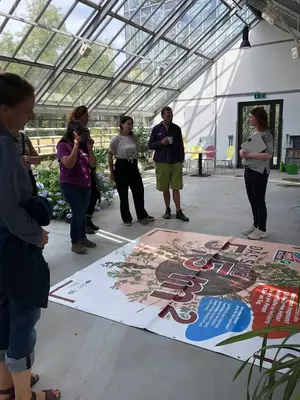 Zu sehen ist eine Gruppe Menschen in einem Innenraum, die um ein großes Plakat herum stehen, das auf dem Boden liegt. Eine Person erklärt etwas. Auf dem Plakat sind Infografiken und -texte zu sehen.