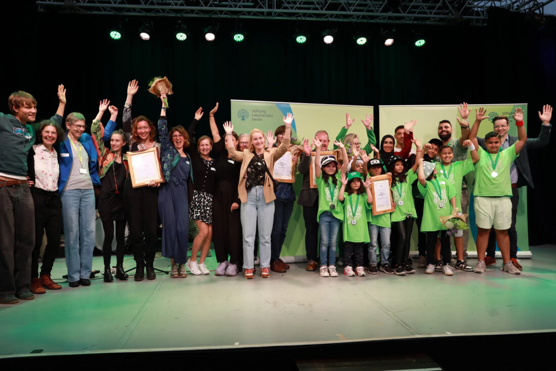 Viele Menschen stehen beisammen und jubeln bei einer Preisverleihung. Sie halten Urkunden oder lächeln in die Kamera. Es handelt sich um die Verleihung des Berliner Naturschutzpreises 2025 durch die Stiftung Naturschutz. Unter den Geehrten sind Weltacker Berlin e.V., Coole Kids Rap, Alexis Tinker-Tsavalas und Frank Sieste.